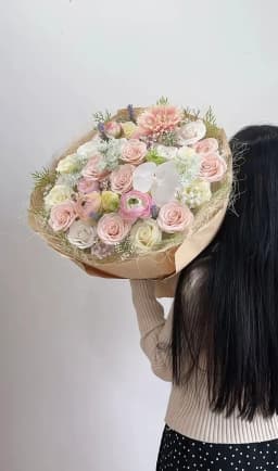 Bó hoa dáng tròn với hồng phấn, hoa trắng kem và vàng nhạt, điểm lan trắng, viền sợi trang trí và giấy gói be.