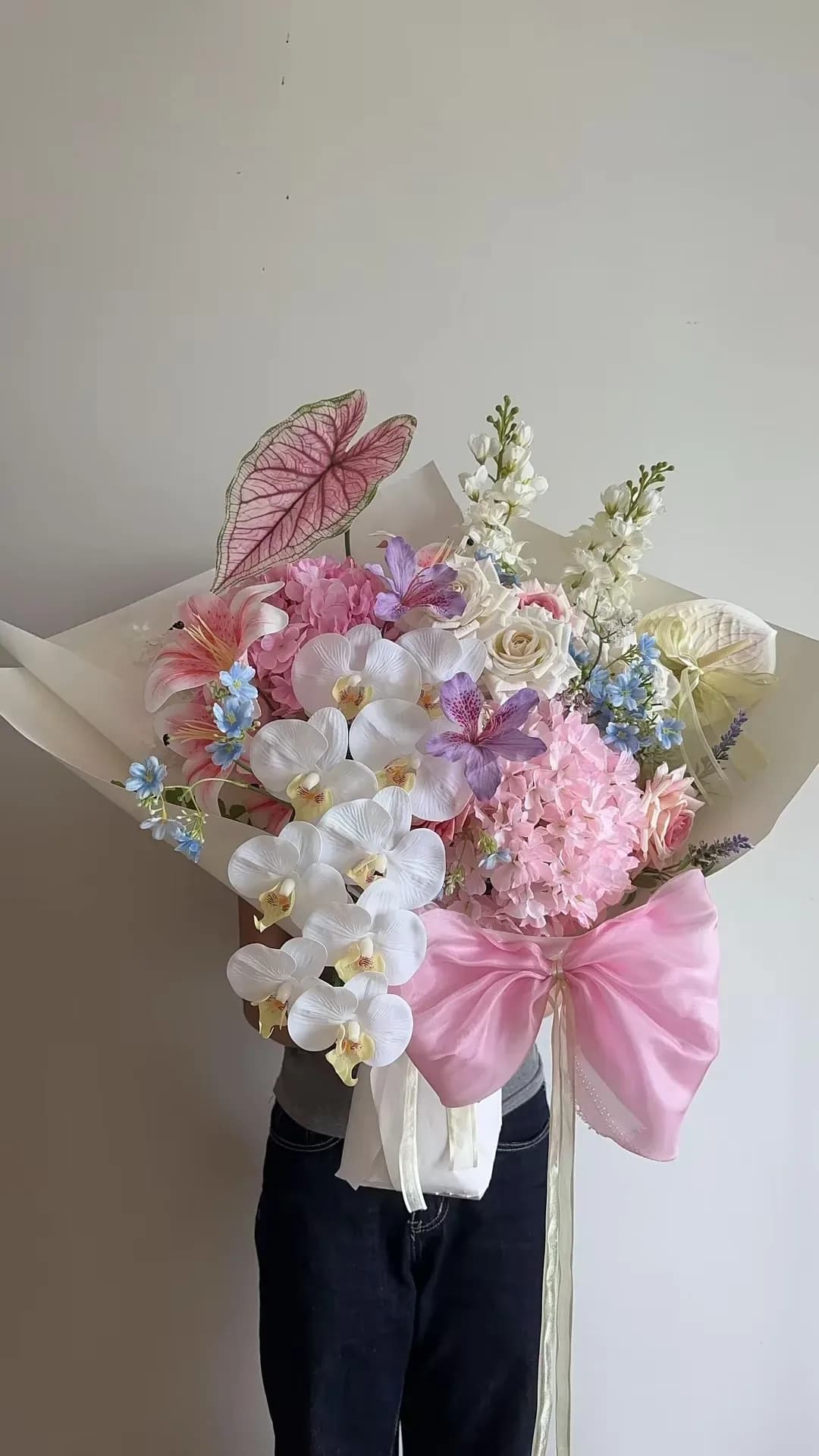 Bó hoa cẩm tú cầu hồng, dải lan trắng rủ phía trước, hoa ly hồng, hoa tím xanh, giấy gói kem và nơ hồng lớn.