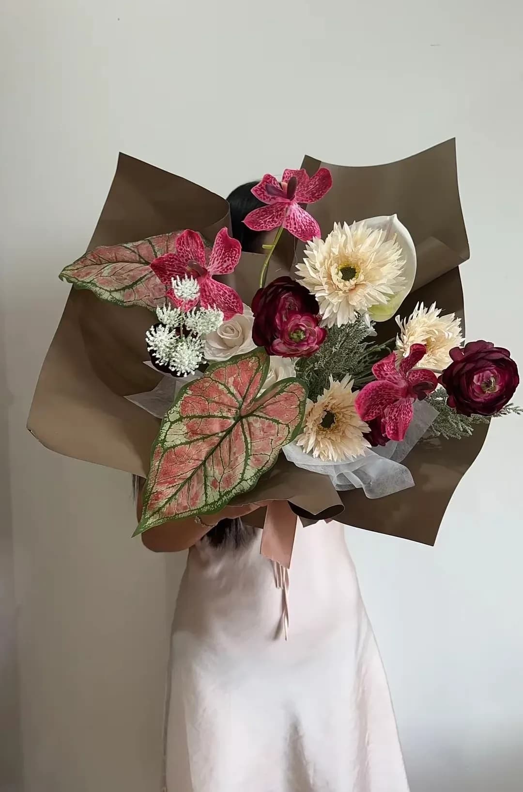 Bó hoa đỏ mận, lan hồng tím và hoa kem, phối lá hồng xanh lớn trong giấy gói nâu, thắt nơ hồng nhạt.