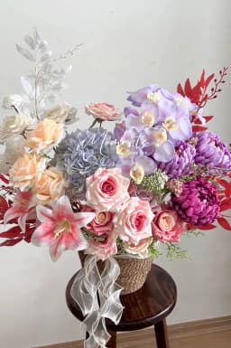 Giỏ cói cắm hoa vải lan hồ điệp tím, cúc tím, cẩm tú cầu xanh, hồng đào và ly hồng, buộc ruy băng voan trắng, đặt trên ghế gỗ – Flowerly Đà Nẵng